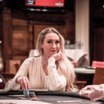Ein Mann in einem schwarzen Anzug und einer roten Krawatte sitzt an einem Pokertisch und hält seine Karten in der Hand. In der Mitte des Tisches liegen Pokerchips und Spielkarten. Im