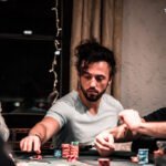 Ein Mann sitzt an einem Pokertisch und hält seine Karten in der Hand, während er konzentriert und ernsthaft auf das Spielgeschehen schaut. Um ihn herum sind Pokerchips und Spielkarten