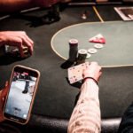 Ein Tablett mit verschiedenen Pokerchips und Spielkarten liegt auf einem grünen Pokertisch. Ein Spieler mit einem Stapel Chips vor sich hält zwei Karten in der Hand und betrachtet sie konzentriert.