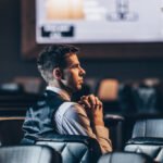 Ein Mann in einem dunkelblauen Anzug und einer roten Krawatte sitzt an einem Pokertisch und betrachtet konzentriert seine Karten. Im Hintergrund sind andere Spieler und Spielchips zu sehen.