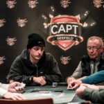 Ein Mann und eine Frau spielen Poker an einem eleganten Tisch mit grüner Filzdecke in einem stilvollen Raum mit Kronleuchter. Der Mann hat gerade Karten ausgeteilt und beide konzentrieren sich