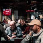 Ein Mann und eine Frau spielen Poker am Tisch, während sie Karten in der Hand halten und konzentriert auf ihre Chips schauen. Die Frau hat blonde Haare und trägt ein rotes Oberteil, während der Mann dunk