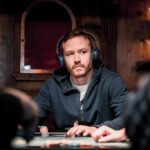 Ein Mann spielt Poker an einem Tisch mit grüner Filzdecke. Er hält Karten in der Hand und schaut konzentriert auf seine Chips. Im Hintergrund sind weitere Spieler und Gläser mit Get