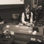 Ein Mann spielt Poker an einem Tisch mit grüner Filzdecke. Er hält Karten in der Hand und betrachtet konzentriert seine Chips. Im Hintergrund sind weitere Spieler und ein Stapel Pokerch