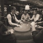 Ein Mann sitzt an einem Pokertisch und hält Karten in der Hand. Er schaut konzentriert auf seine Karten, während andere Spieler im Hintergrund zu sehen sind. Der Tisch ist mit Chips und Getr