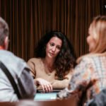 Ein Mann in einem eleganten Anzug sitzt an einem Pokertisch und hält seine Hand mit Spielkarten. Im Hintergrund sind weitere Spieler und Spielchips zu sehen.