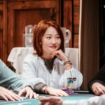 Ein Tisch mit grünem Pokertuch, auf dem Pokerchips und Spielkarten liegen. Vier Personen sitzen um den Tisch herum und spielen Poker.