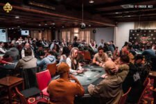 Ein entspannter Mann sitzt an einem Pokertisch mit einem Stapel Pokerchips vor sich. Er hält zwei Spielkarten in der Hand und schaut konzentriert auf den Tisch. Im Hintergrund sind