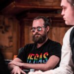 Ein Mann sitzt an einem Pokertisch mit Chips und Karten. Er betrachtet nachdenklich seine Hand, während er von anderen Spielern beobachtet wird.