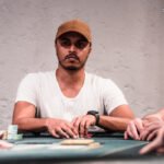 Ein Mann sitzt an einem Pokertisch und hält zwei Spielkarten in der Hand, während er konzentriert auf die Karten auf dem Tisch schaut. Andere Spieler sind im Hintergrund zu sehen.