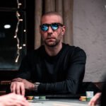 Ein bunter Pokertisch mit Spielkarten, Pokerchips und Getränken, umgeben von Menschen, die konzentriert spielen.