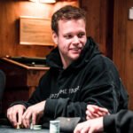 Ein Mann sitzt an einem Pokertisch und hält zwei Karten in der Hand. Er schaut konzentriert auf die Mitte des Tisches, wo Pokerchips und Spielkarten liegen. Im Hintergrund sind