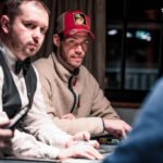 Ein professioneller Pokertisch mit grünem Filz, umgeben von Stühlen, Chips und Spielkarten. Vier Spieler sitzen an dem Tisch und konzentrieren sich auf ihre Karten. Helle Bele