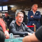 Ein Mann in einem schwarzen Anzug und einer roten Krawatte sitzt an einem Pokertisch und hält seine Karten in der Hand. Er schaut konzentriert auf den Tisch, während im Hintergrund andere