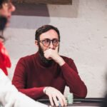 Ein Mann in einem schwarzen Anzug und einer dunkelblauen Krawatte sitzt an einem Pokertisch und hält seine Spielkarten in der Hand. Er schaut konzentriert auf den Tisch. Im Hinter
