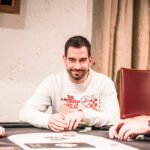 Ein professioneller Pokertisch mit grünem Filz, umgeben von Stühlen und Chips. In der Mitte des Tisches liegen Spielkarten und eine Hand hält zwei Asse. Im Hintergrund sind unschar