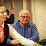 Ein Mann sitzt an einem Pokertisch und hält Karten in der Hand, während er konzentriert auf das Spiel schaut. Um ihn herum sind weitere Spieler zu sehen, die ebenfalls Karten halten.
