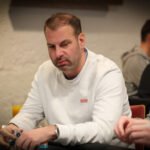 Ein Mann sitzt an einem Pokertisch und hält seine Karten in der Hand, während er konzentriert auf den Tisch schaut. Im Hintergrund sind weitere Spieler und ein Stapel Pokerchips zu sehen.