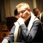 Ein Mann in einem schwarzen Anzug und einer hellen Krawatte sitzt an einem Pokertisch, während er konzentriert seine Karten betrachtet. Um ihn herum sind Pokerchips und ein Kartendeck verte