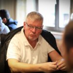 Ein Mann sitzt an einem Pokertisch und hält zwei Karten in der Hand. Er schaut konzentriert auf die Gemeinschaftskarten in der Mitte des Tisches. Im Hintergrund sind andere Spieler und Poker