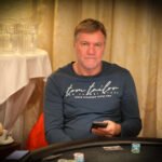 Ein Mann in einem dunkelblauen Anzug und einer schwarzen Krawatte sitzt an einem Pokertisch und hält eine Hand mit Spielkarten. Im Hintergrund sind weitere Spieler und Pokerchips zu sehen.