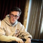 Ein Mann in einem schwarzen Anzug und einer schwarzen Sonnenbrille spielt Poker an einem eleganten Tisch in einem Casino, umgeben von anderen Spielern.