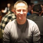 Ein Mann mit einem Pokerchip-Stapel vor sich sitzt an einem Pokertisch, umgeben von anderen Spielern. Er betrachtet seine Karten mit ernstem Gesichtsausdruck.