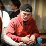 Ein Mann spielt Poker an einem Tisch in einem eleganten Casino. Er hat eine ernste Miene und hält seine Karten in der Hand, während er aufmerksam auf den Tisch schaut. Im Hintergrund sind andere