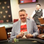 Ein professioneller Pokerspieler mit Sonnenbrille und Kappe, der konzentriert Karten auf einem grünen Pokertisch betrachtet. Im Hintergrund sind weitere Spieler und Chips zu sehen.