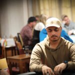 Ein Mann sitzt an einem Pokertisch und hält seine Karten mit ernstem Gesichtsausdruck. In der Mitte des Tisches liegen Pokerchips und Spielkarten. Im Hintergrund sind weitere Personen uns