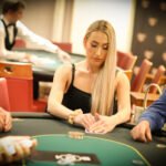 Ein Mann sitzt an einem Pokertisch und hält zwei Spielkarten in der Hand, während er konzentriert auf die Gemeinschaftskarten auf dem Tisch schaut. Andere Spieler und ein Stapel Pokerchips