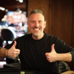 Ein Mann in einem schwarzen Anzug und einer Fliege sitzt an einem Pokertisch und hält eine Hand mit Spielkarten. Er hat eine ernste Miene und scheint konzentriert zu sein. Im Hinter