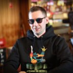 Ein Mann sitzt an einem Pokertisch und hält zwei Spielkarten in der Hand. Er trägt ein schwarzes Sakko und eine dunkle Sonnenbrille. Im Hintergrund sind weitere Pokertische und Spieler zu sehen