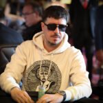 Ein Mann sitzt an einem Pokertisch und hält Karten in der Hand, während er konzentriert auf seine Gegner schaut. Die anderen Spieler im Hintergrund sind unscharf zu sehen.