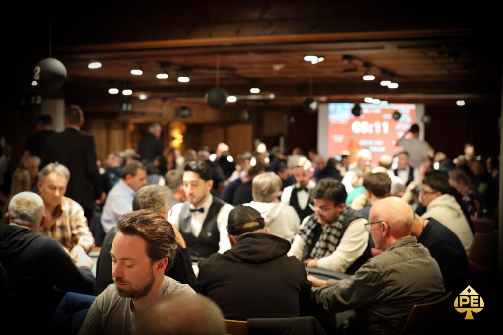 Ein Mann und eine Frau sitzen an einem Pokertisch und betrachten konzentriert ihre Karten. Die Frau hat blonde Haare und trägt eine schwarze Bluse, der Mann eine Brille und ein graues Hemd.