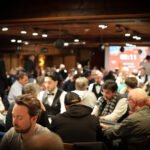 Ein Mann und eine Frau sitzen an einem Pokertisch und betrachten konzentriert ihre Karten. Die Frau hat blonde Haare und trägt eine schwarze Bluse, der Mann eine Brille und ein graues Hemd.