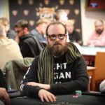 Ein Mann sitzt an einem Pokertisch und hält eine Hand mit Spielkarten. Er trägt einen dunklen Anzug und eine schwarze Krawatte. Um ihn herum sind Pokerchips und Getränke auf dem