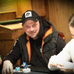 Ein Mann in einem dunkelblauen Anzug und einer hellblauen Krawatte sitzt an einem Pokertisch und betrachtet konzentriert seine Karten. Im Hintergrund sind Pokerchips und eine Glühlam