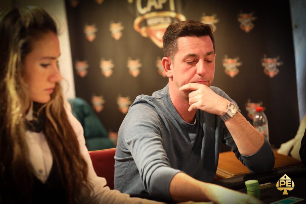 Ein professioneller Pokerspieler sitzt an einem Tisch mit Pokerchips vor sich. Er hält seine Karten in der Hand und schaut konzentriert auf den Tisch. Im Hintergrund sind weitere Spieler und eine