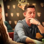 Ein professioneller Pokerspieler sitzt an einem Tisch mit Pokerchips vor sich. Er hält seine Karten in der Hand und schaut konzentriert auf den Tisch. Im Hintergrund sind weitere Spieler und eine
