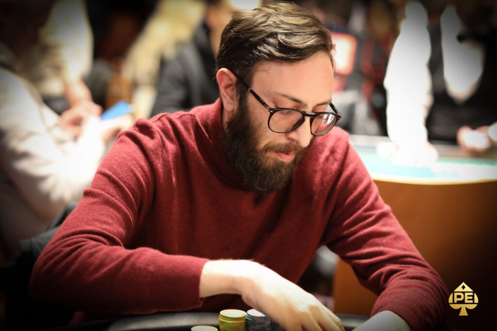 Ein Mann spielt Poker an einem Tisch in einem eleganten Casino. Er hält seine Karten in der Hand und schaut konzentriert auf den Tisch. Um ihn herum sitzen andere Spieler und beobachten das Spiel