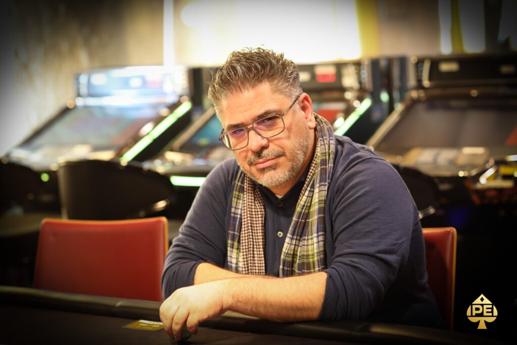 Ein Mann mit einer schwarzen Mütze und einem grauen Pullover sitzt an einem Pokertisch und hält seine Karten. Im Hintergrund sind weitere Spieler und Pokerchips zu sehen.