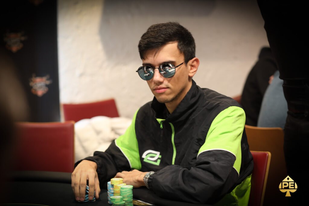 Ein Mann mit schwarzer Sonnenbrille und rotem Anzug sitzt lässig an einem Pokertisch und hält seine Karten in der Hand. Im Hintergrund sind weitere Spieler und Casino-Chips zu sehen.