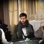 Ein Mann in einem dunkelblauen Anzug und einer weißen Hemdweste spielt Poker an einem eleganten Holztisch in einem luxuriösen Casino. Er hält seine Karten in der Hand und betrachtet sie
