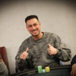 Ein Mann sitzt an einem Pokertisch mit grünem Filz und hält zwei Spielkarten in der Hand. Er scheint konzentriert auf seine Hand zu schauen, während andere Spieler im Hintergrund verschwommen