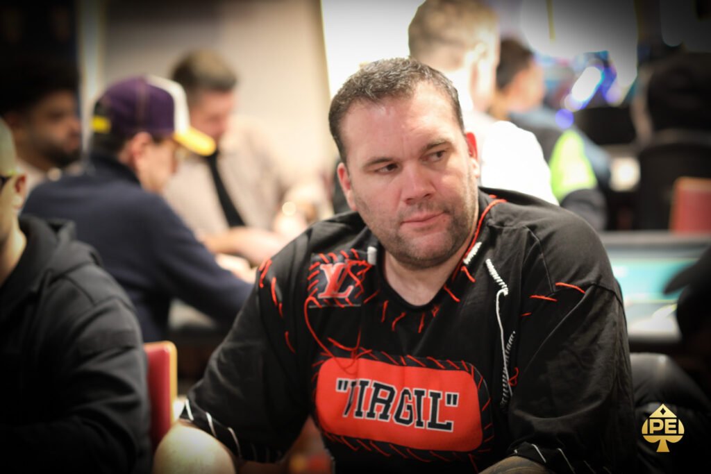 Ein Mann in einem schwarzen Anzug und schwarzen Hut sitzt an einem Pokertisch und hält seine Karten in der Hand, während er konzentriert auf das Spiel schaut.
