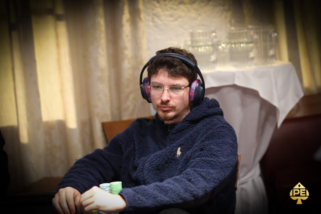 Ein Mann sitzt an einem Pokertisch und hält seine Karten mit ernstem Gesichtsausdruck. Um ihn herum sind andere Spieler und Jetons zu sehen.