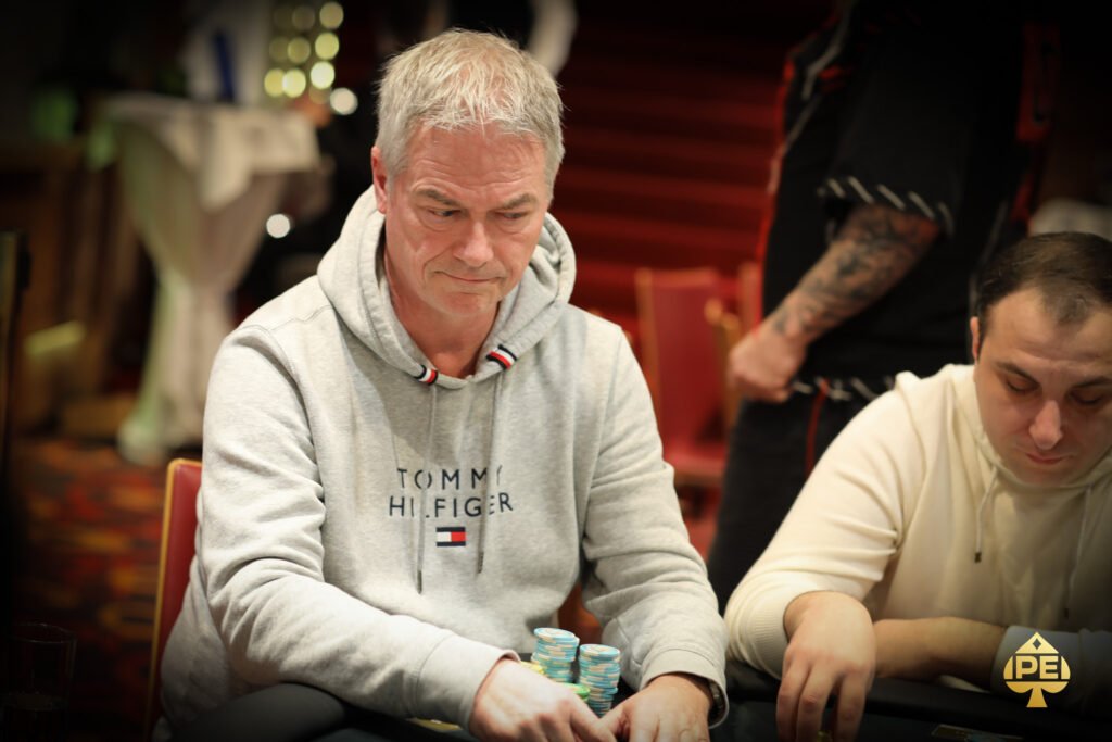 Ein Mann sitzt an einem Pokertisch und hält eine Hand mit Spielkarten. Er hat eine ernsthafte Miene und trägt ein dunkles Sakko. Andere Spieler sind im Hintergrund zu sehen, die ihre