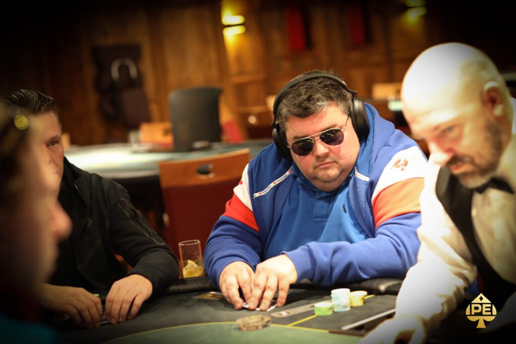 Ein Mann sitzt an einem Pokertisch und hält zwei Spielkarten in der Hand. Er schaut konzentriert auf den Tisch. Im Hintergrund sind andere Spieler und Pokerchips zu sehen.