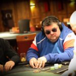 Ein Mann sitzt an einem Pokertisch und hält zwei Spielkarten in der Hand. Er schaut konzentriert auf den Tisch. Im Hintergrund sind andere Spieler und Pokerchips zu sehen.